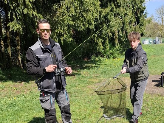 Zwei Männer stehen auf einer grünen Wiese vor hohen Nadelbäumen. Der Mann links hält eine Angelrute, während der Mann rechts ein Kescher mit einem Fisch hält. Beide tragen funktionelle Outdoor-Kleidung.