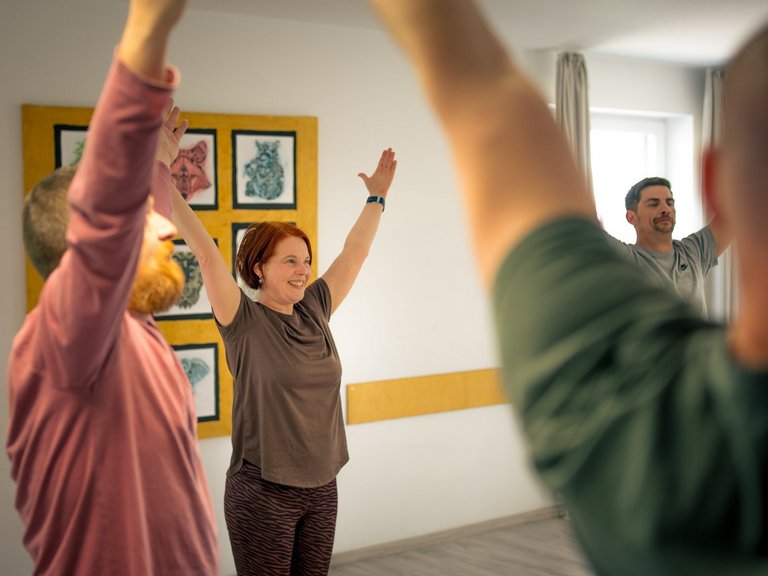 Eine lächelnde Frau mit roten Haaren steht im Mittelpunkt einer Gruppe und hebt beide Arme nach oben. Links und rechts von ihr sind weitere Personen zu sehen, die die gleiche Bewegung ausführen, vermutlich bei einer Yoga- oder Gymnastikstunde in einem hellen Raum.