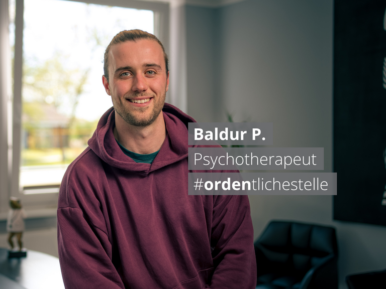 Ein lächelnder junger Mann in einem burgunderroten Hoodie sitzt vor einem Fenster. Ein Textfeld neben ihm zeigt: „Baldur P. Psychotherapeut #ordentlichestelle".